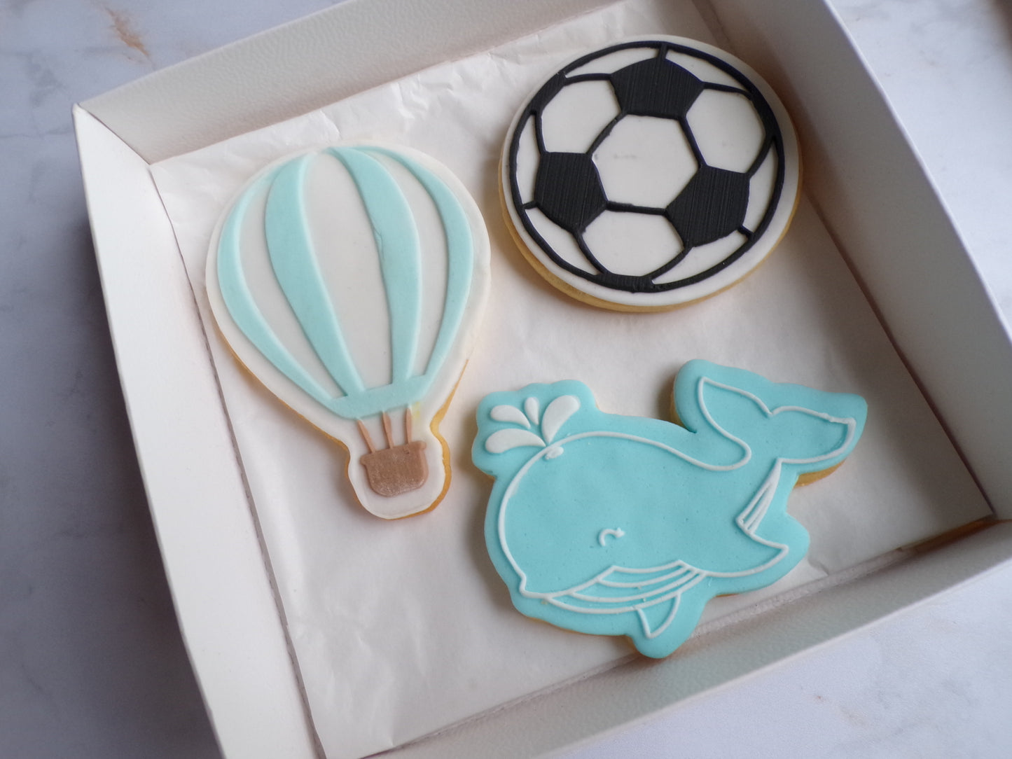 Boîte contenant trois biscuits sablés décorés pour enfant ou naissance : un biscuit en forme de montgolfière bleu pastel et blanc avec un panier marron, un biscuit rond décoré comme un ballon de football noir et blanc, et un biscuit en forme de baleine bleue souriante avec un jet d'eau. Biscuits décorés à la main, thème naissance ou baby shower garçon.