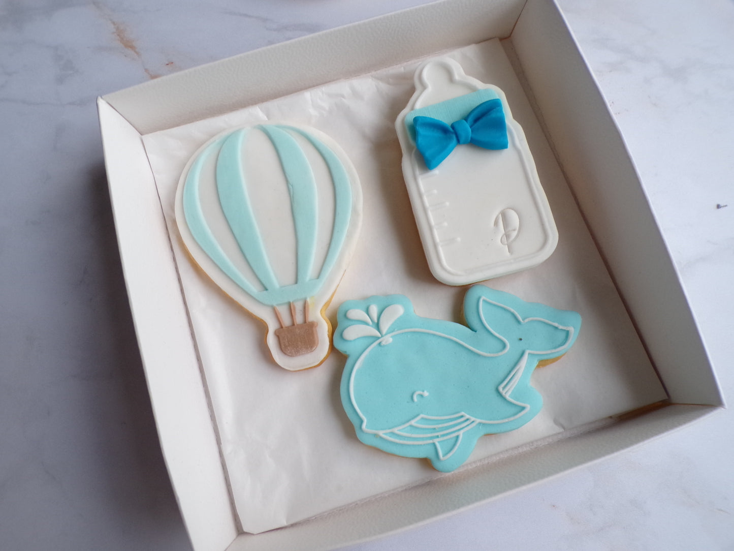 Boîte contenant trois biscuits sablés décorés pour enfant : un biscuit en forme de montgolfière bleu pastel et blanc avec un panier marron, un biscuit en forme de biberon blanc avec un noeud papillon bleu, et un biscuit en forme de baleine bleue souriante avec un jet d'eau. Biscuits décorés à la main, thème naissance ou baby shower garçon.