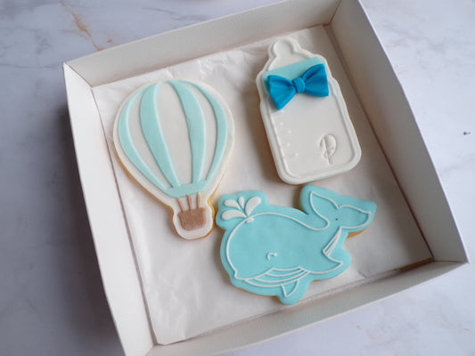 Boîte contenant trois biscuits sablés décorés pour enfant : un biscuit en forme de montgolfière bleu pastel et blanc avec un panier marron, un biscuit en forme de biberon blanc avec un noeud papillon bleu, et un biscuit en forme de baleine bleue souriante avec un jet d'eau. Biscuits décorés à la main, thème naissance ou baby shower garçon.