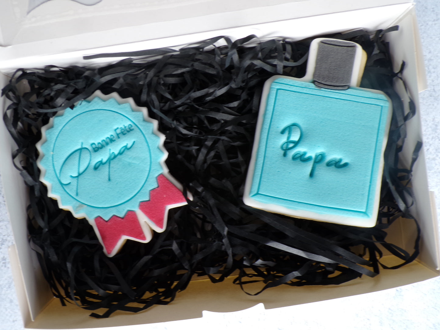 coffret de deux biscuits décorés en pâte à sucre. il y a un biscuit en forme de médaille bleu et rouge avec l'inscription "bonne fête papa" et un autre biscuit en forme de parfum bleu et le bouchon gris avec la mention papa au centre