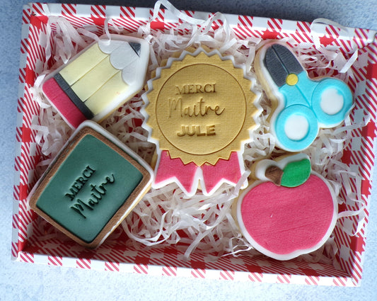 Box composé de 5 biscuits sablés décoré en pâte à sucre. il y a un biscuit en forme de tablea avec l'inscription "merci maitre", un biscuits en forme de ciseau bleu et noir, un biscuits en forme de médaille or et rouge avec l'inscription " merci maitre jule" un biscuit en forme de pomme rouge et un biscuit en forme de crayon.