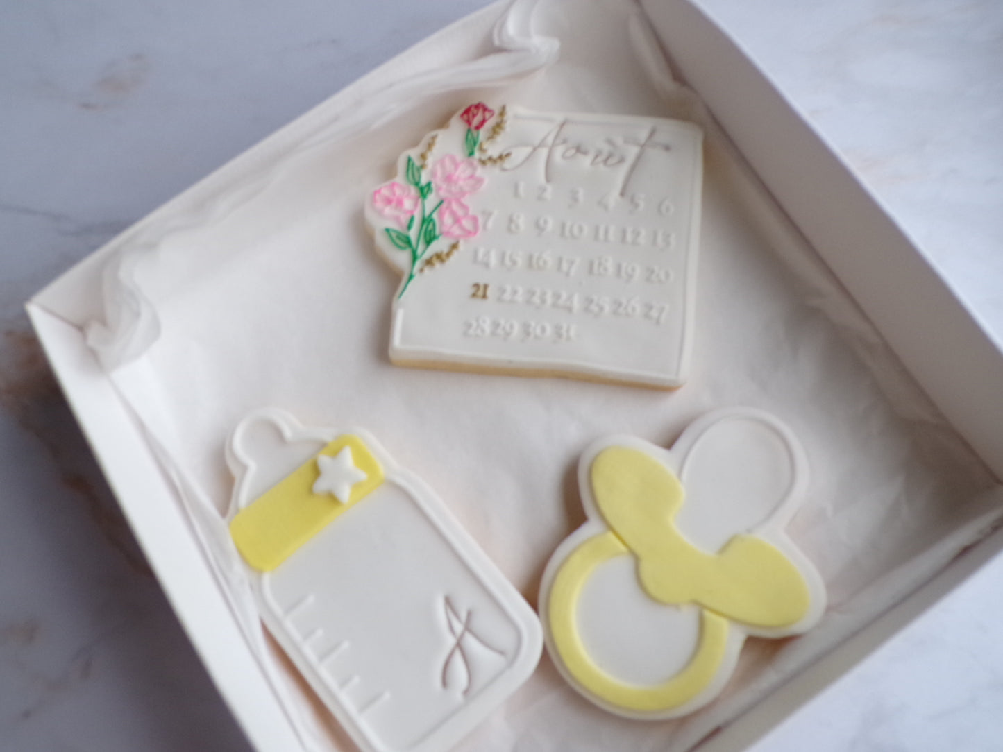 Boîte contenant trois biscuits sablés décorés pour enfant : un biscuit en forme de calendrier avec une fleur sur le côté, un biscuits en forme de biberon blanc et j'aune avec une étoile blanche dessus et un biscuits en forme de tétine jaune et blanc