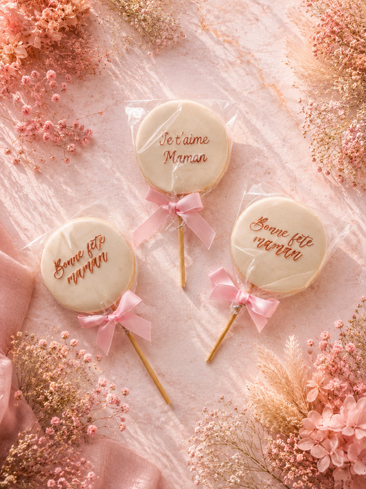 Sucettes biscuits artisanales décorées en pâte à sucre avec messages Fête des Mères et rubans roses sur fond floral rose poudré