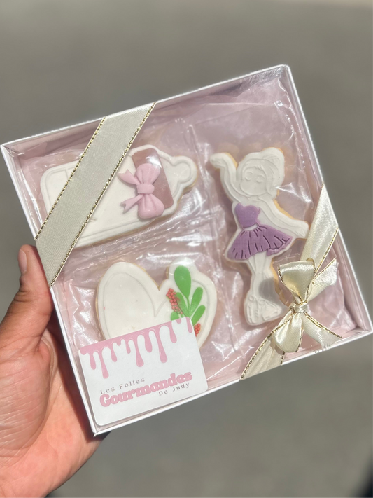 Boite avec un ruban beige et les bords doré. la boite contenant trois biscuits sablés décorés : un biscuits en forme de biberon blanc et un noeud papillon rose claire, un biscuits en forme de danseuse blanche avec une robe violet et un biscuits en forme de coeur blanc une feuille verte avec un petite boule rouge