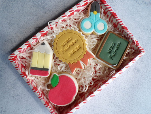 Box composé de 5 biscuits sablés décoré en pâte à sucre. il y a un biscuit en forme de tablea avec l'inscription "merci maitresse", un biscuits en forme de ciseau bleu et noir, un biscuits en forme de médaille or et rouge avec l'inscription " merci maitresse julie" un biscuit en forme de pomme rouge et un biscuit en forme de crayon.